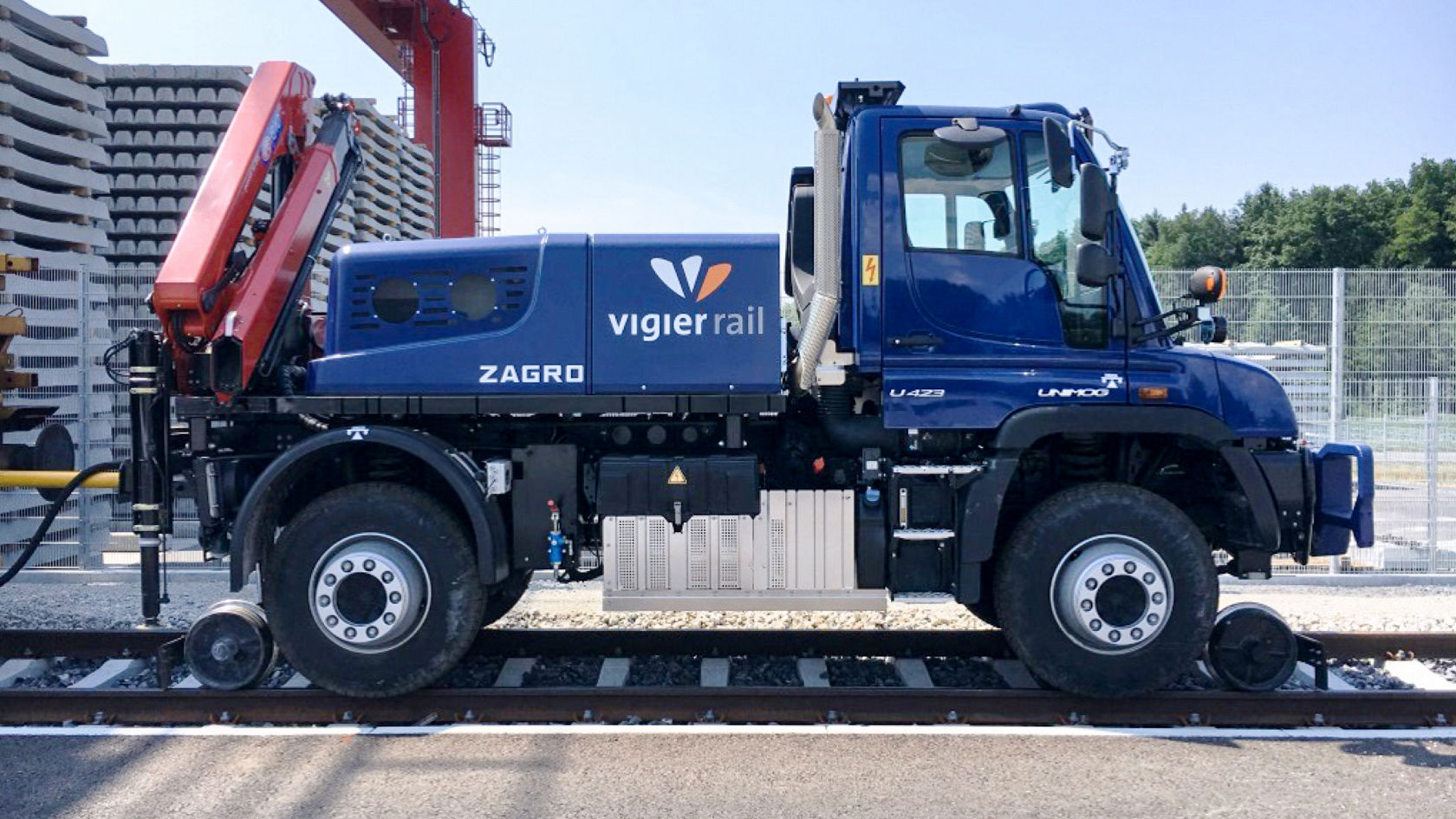蓝色 Unimog 带铁路设备和字标，停放在工业场地的轨道上。